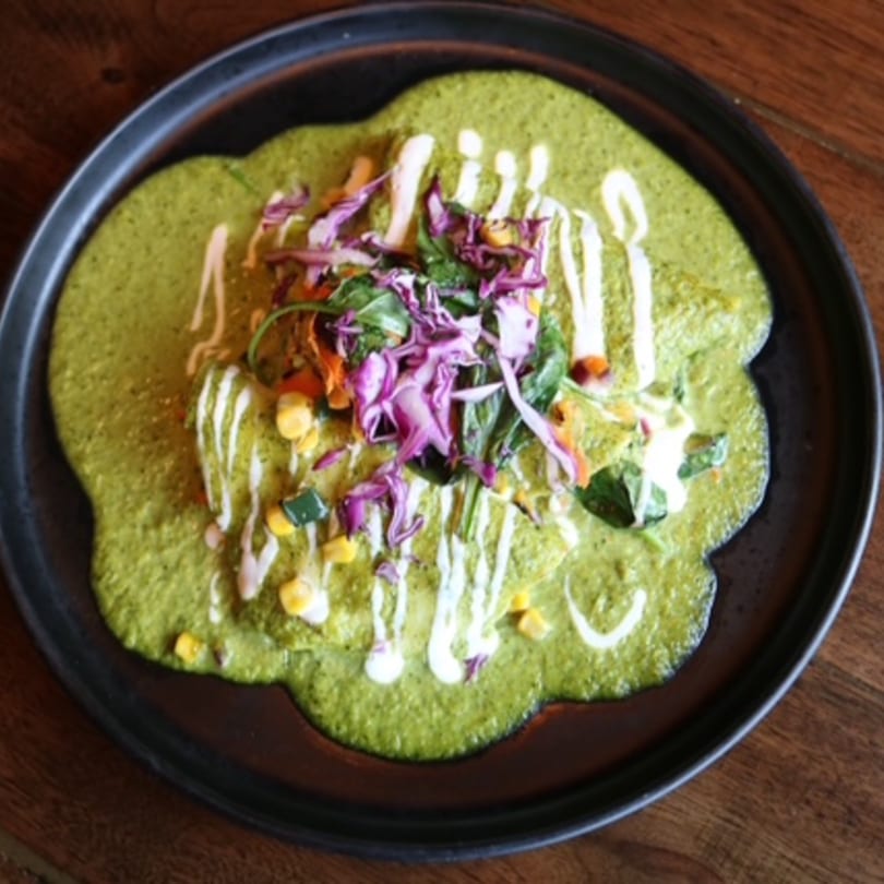 Green enchiladas with white crema, purple cabbage, corn, and other vegetables on a black plate.