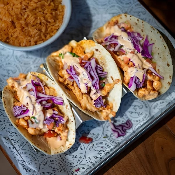 Three chicken tacos with purple cabbage, sauce, and a side of rice on a metal tray.