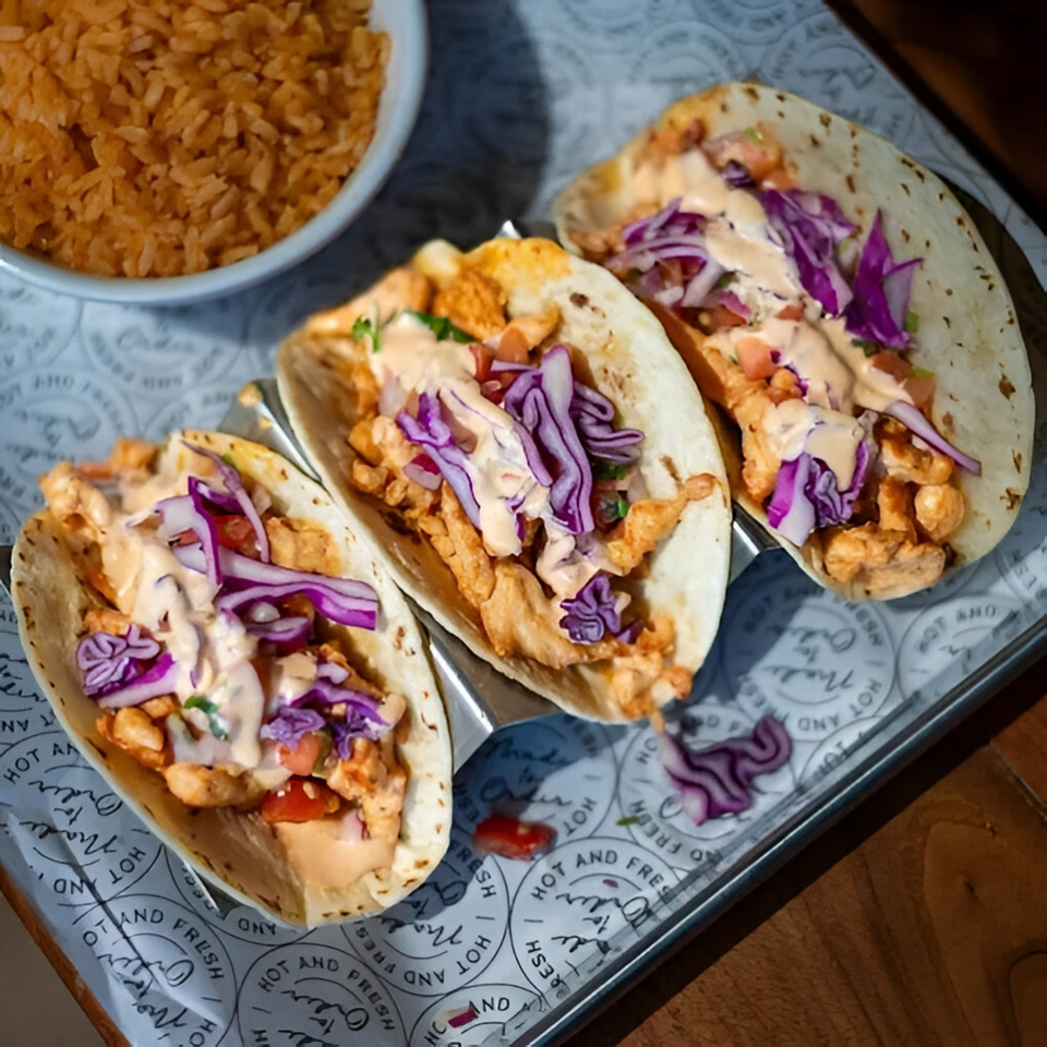 Three chicken tacos with purple cabbage, sauce, and a side of rice on a metal tray.